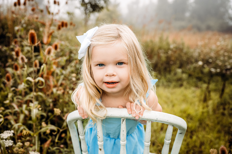 Family Photography, Little girl holding onto the back of an old white chair, looking at camera